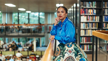 Terezia is leaning against a wooden balcony rail overlooking the work area of a library