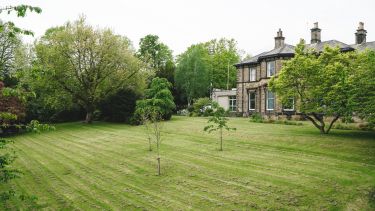 A photograph of a large green out door space, with trees, and a large Victorian building in the background
